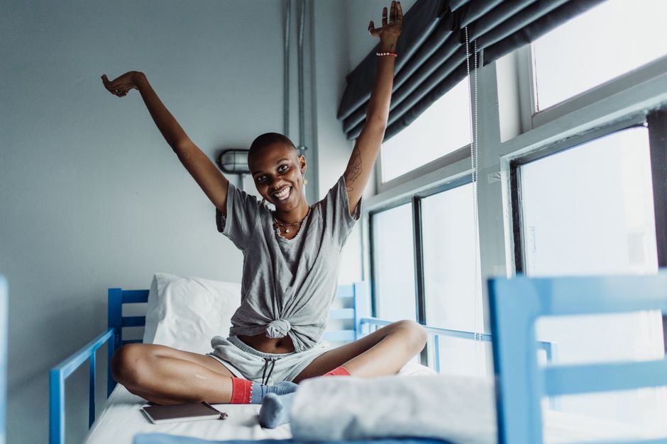 Smiling woman with arms raised sitting on a bunk bed in a bright room.
