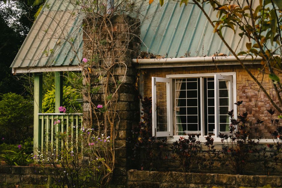 Rustic cottage with open windows and lush garden in a serene setting.