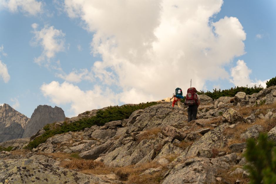 Two hikers with backpacks trek a rocky path in the scenic mountains of Wyoming under a cloudy sky.