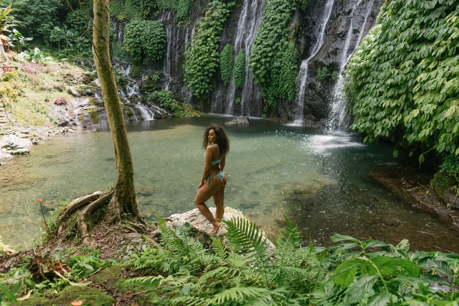 Side view of graceful young ethnic female traveler with curly hair in bikini enjoying nature near clear lake and waterfall in tropical jungle on sunny day