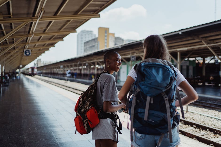 Two diverse female backpackers wait on a sunny railway platform, ready for adventure.