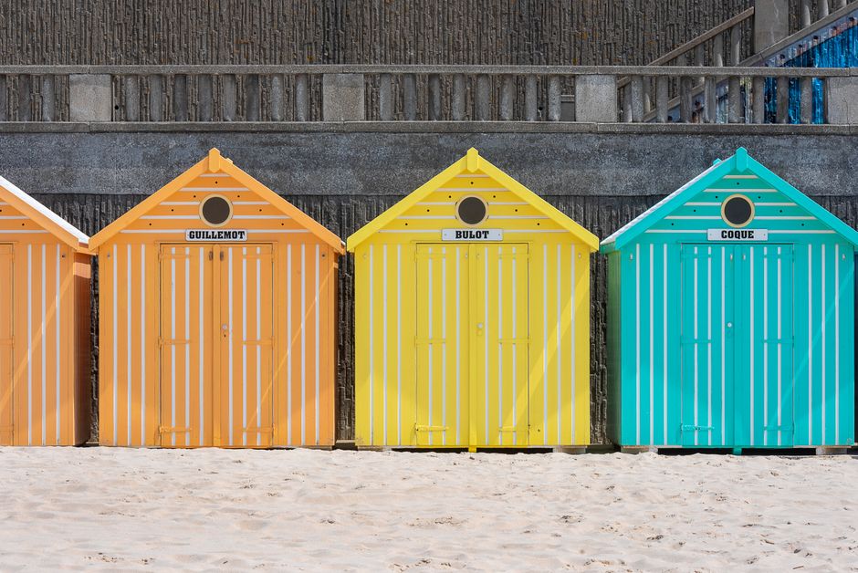 Bright beach huts in orange, yellow, and teal on a sandy beach against a rustic wall.