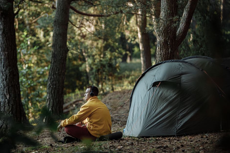 A person enjoying a calm morning by a tent in a forest wearing headphones.
