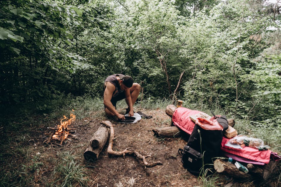 Man setting up campfire in forest with gear spread out. Perfect for outdoor adventure themes.