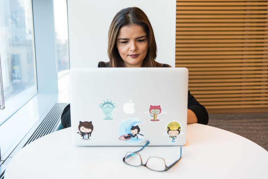 A woman focuses on her laptop adorned with tech-themed stickers, sitting in a bright office.