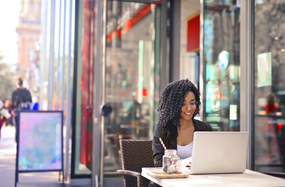 Smiling woman with curly afro hair typing on a laptop outside a modern cafe.