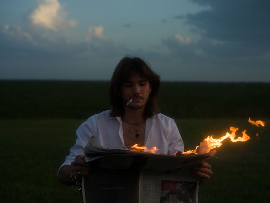 A man with long hair reading a newspaper on fire outdoors at dusk, evoking drama.