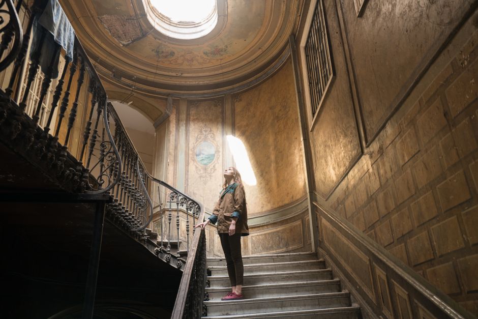 Woman standing on vintage staircase, gazing at ornate ceiling in historic building.