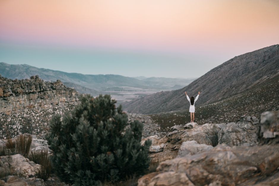 A person stands on rocky terrain with arms raised, embracing the scenic mountain view at sunset.