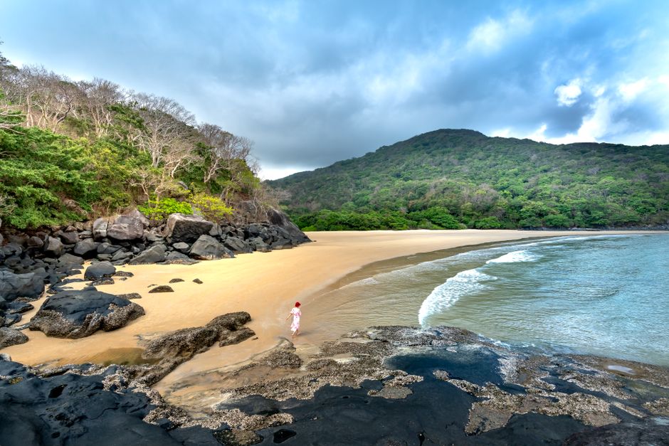 A tranquil beach setting with rocky formations and lush forests under a cloudy sky.