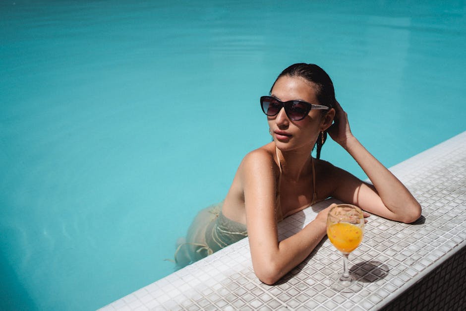 High angle of self esteem young ethnic lady in swimwear and sunglasses touching wet hair while chilling in pool water with glass of fresh orange juice