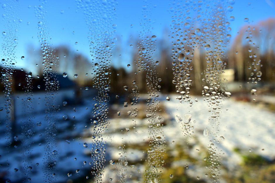 A window with water droplets overlooking a snowy winter landscape under a clear blue sky.