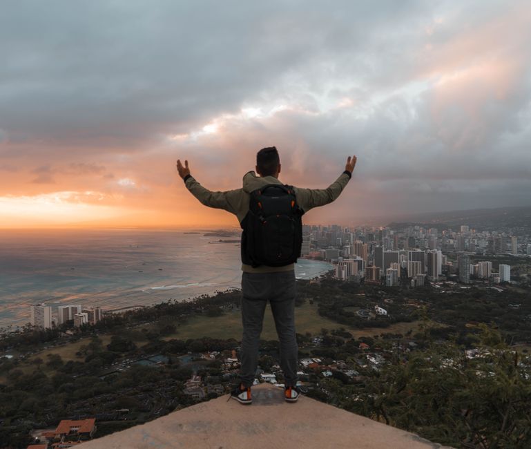 Back view full body male tourist with backpack standing with arms outstretched on hilltop and enjoying majestic view of modern coastal city on overcast day