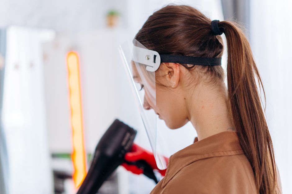A hairdresser wearing a face shield uses a blow dryer in a salon, embracing safety measures.