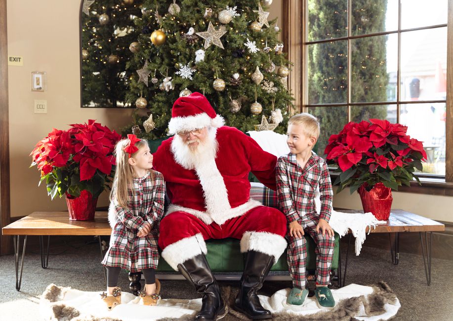 Santa Claus sitting with children by a Christmas tree, surrounded by festive decor and red poinsettias.