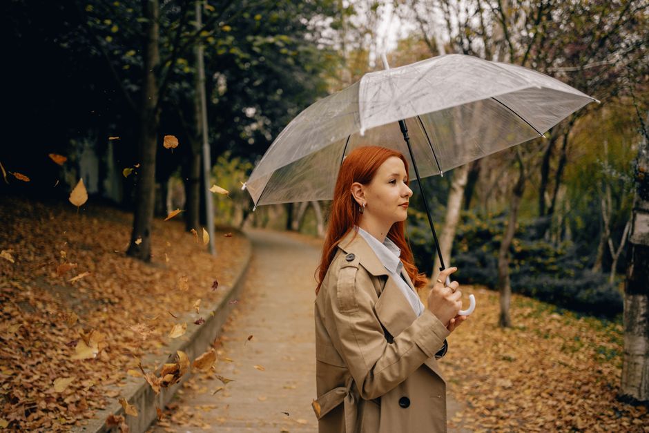 Stylish woman with red hair holding a transparent umbrella in a tranquil autumn park setting.