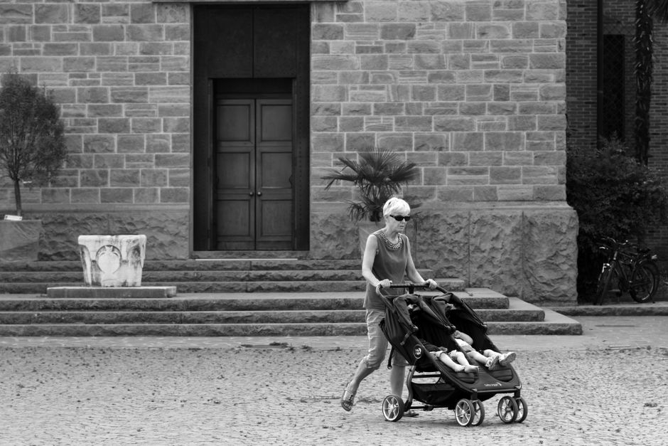 An elderly woman pushes a double stroller in a serene outdoor setting in Abano Terme, Italy.