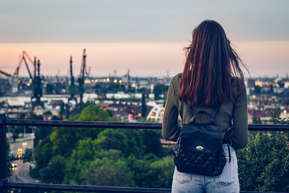 Woman with backpack gazing at Gdańsk's industrial skyline at sunset reflecting urban exploration.