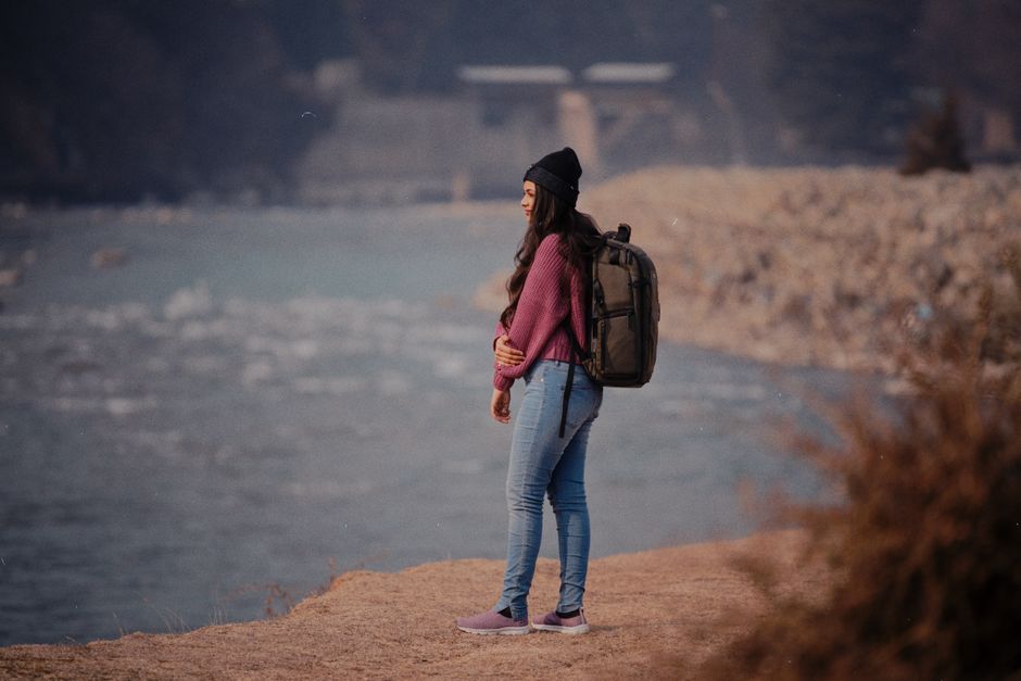 A woman with a backpack hiking along a scenic riverbank, dressed for cool weather.