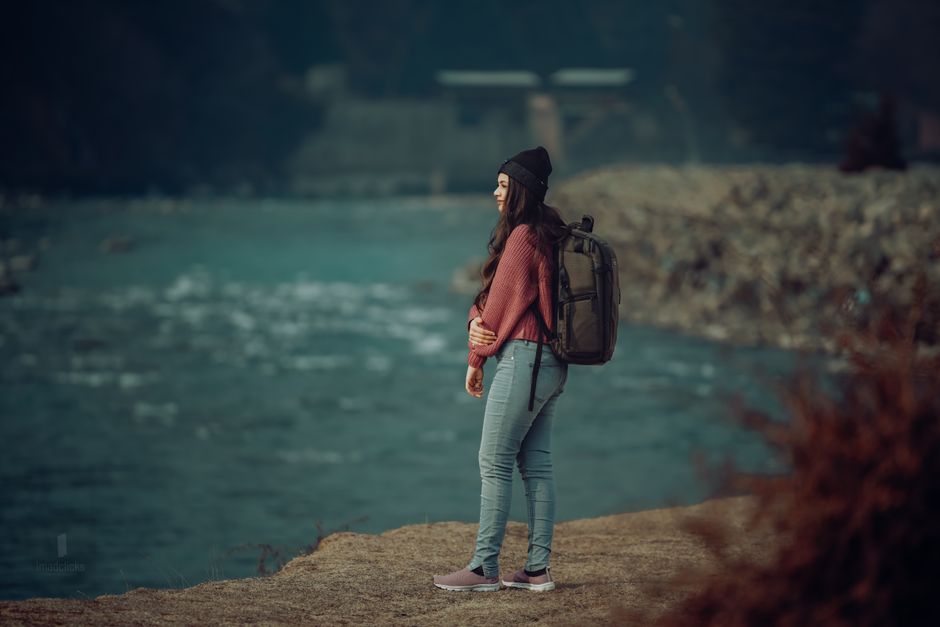A young woman stands by a scenic riverbank wearing a backpack and hat, enjoying the outdoor view.