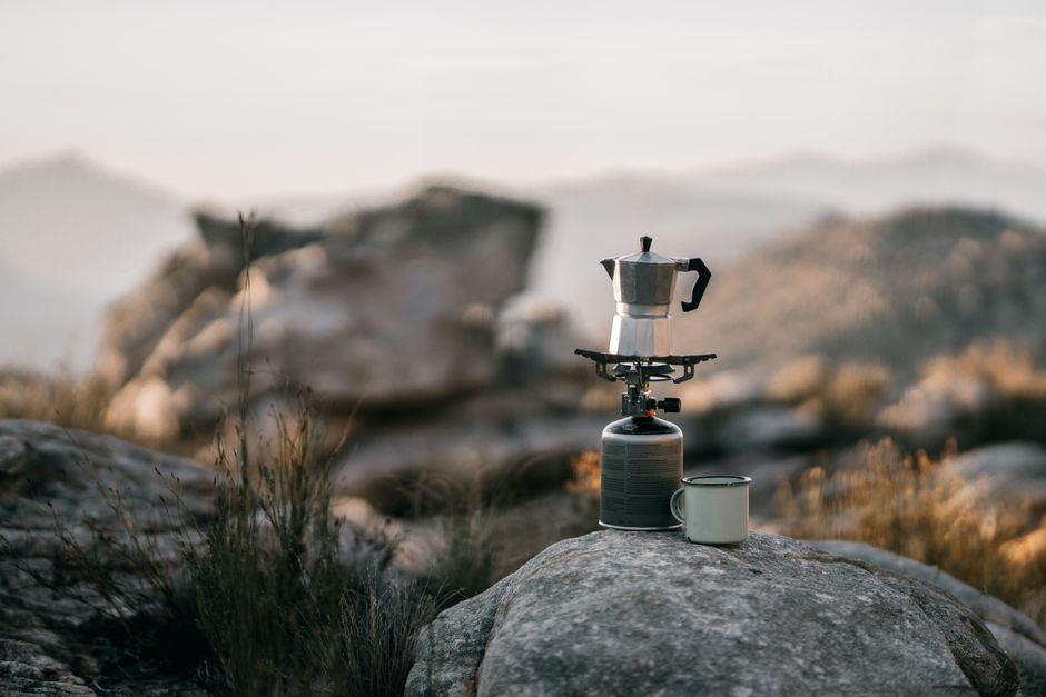 Moka pot and mug on rocks in serene mountain setting, perfect for adventure coffee lovers.