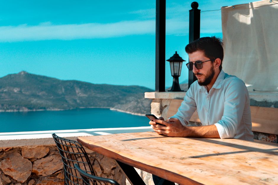 A man using a cellphone while sitting at an outdoor cafe in Antalya, Türkiye, with a mountain view.