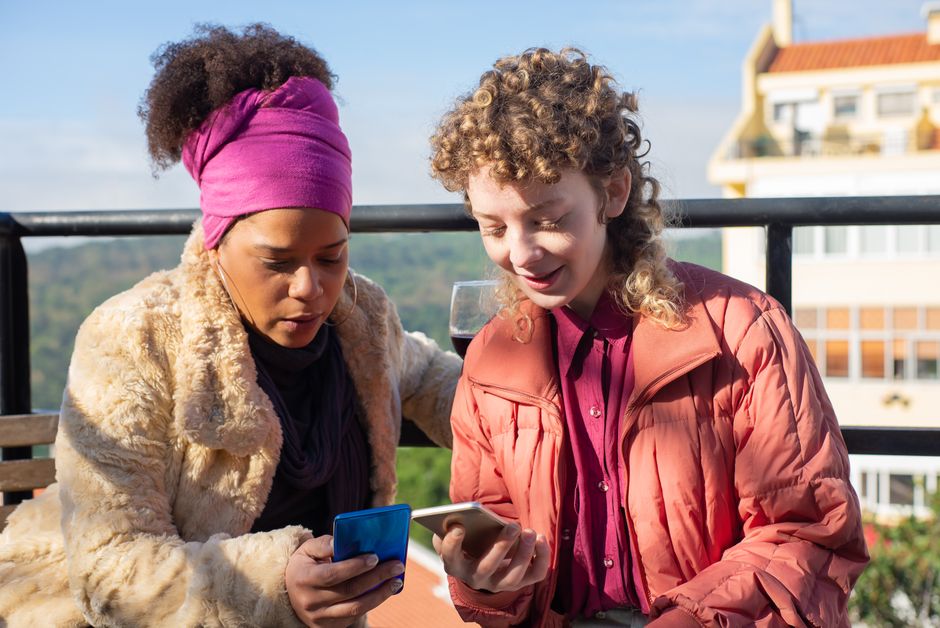 Two women sitting on a rooftop, enjoying wine and using smartphones together.