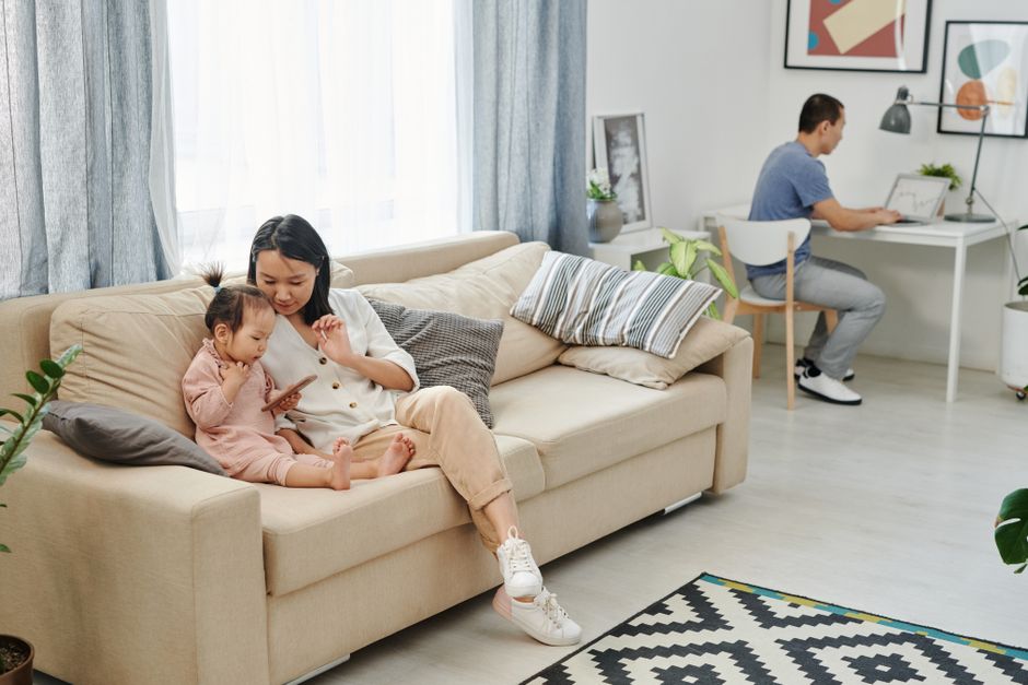 A mother and child bonding on a sofa while a man works on a laptop in a well-lit living room.