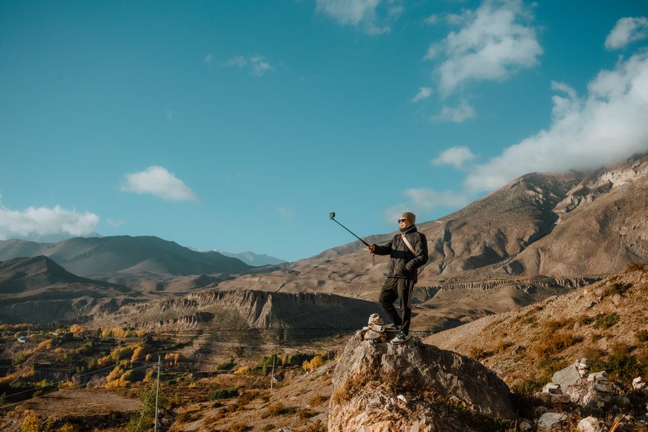 A lone traveler captures a selfie on a rocky peak amidst stunning mountain scenery under a bright blue sky.