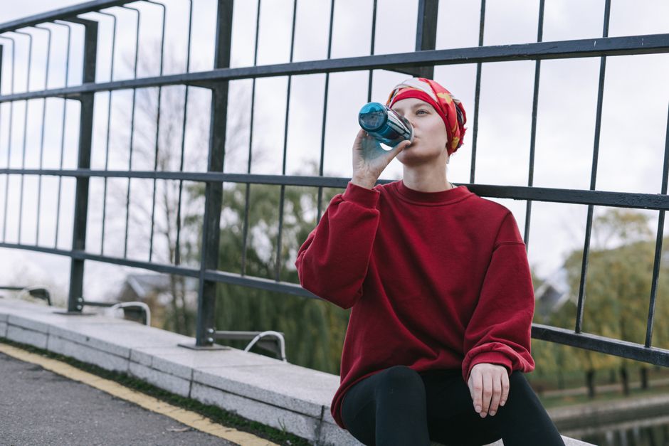 Woman wearing a headscarf and red sweater drinking from a bottle outdoors.