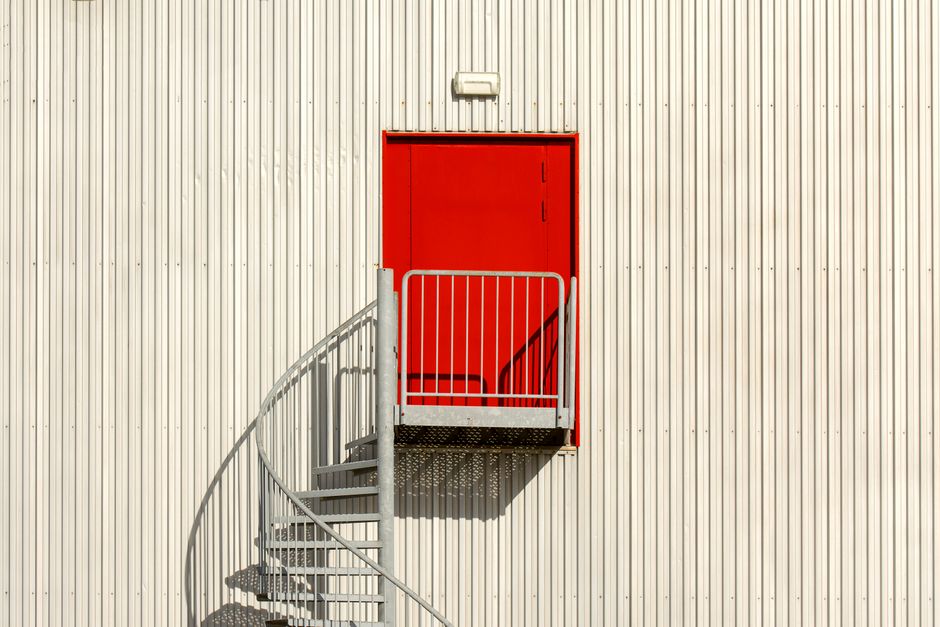 Minimalist architecture with a spiral staircase and vibrant red door against a corrugated metal wall.