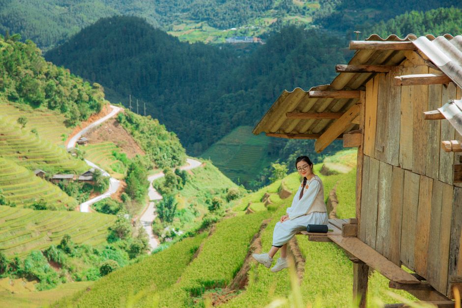 A woman sits by a rustic hut, overlooking lush green terraces and mountains, capturing a serene summer landscape.
