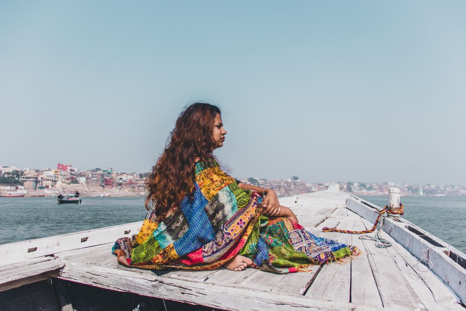 Side view of calm young Indian lady in colorful maxi dress sitting on wooden deck of old boat floating on sea on sunny day