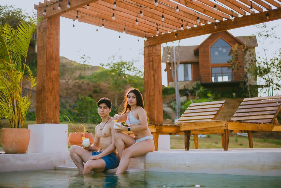 Young couple enjoying drinks by the poolside under a rustic pergola on a sunny day.