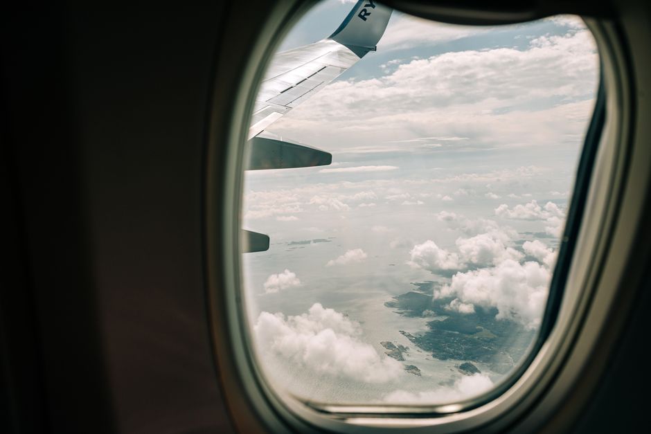 View through airplane window showing wing and ocean with clouds below during daylight flight