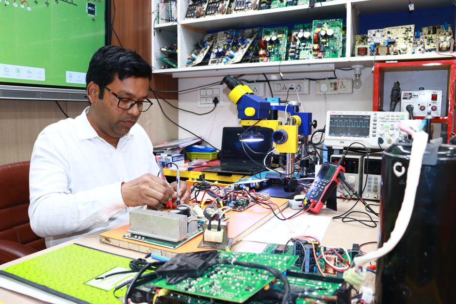 An electronics engineer working on circuit board repairs in a tech workshop filled with tools and equipment.